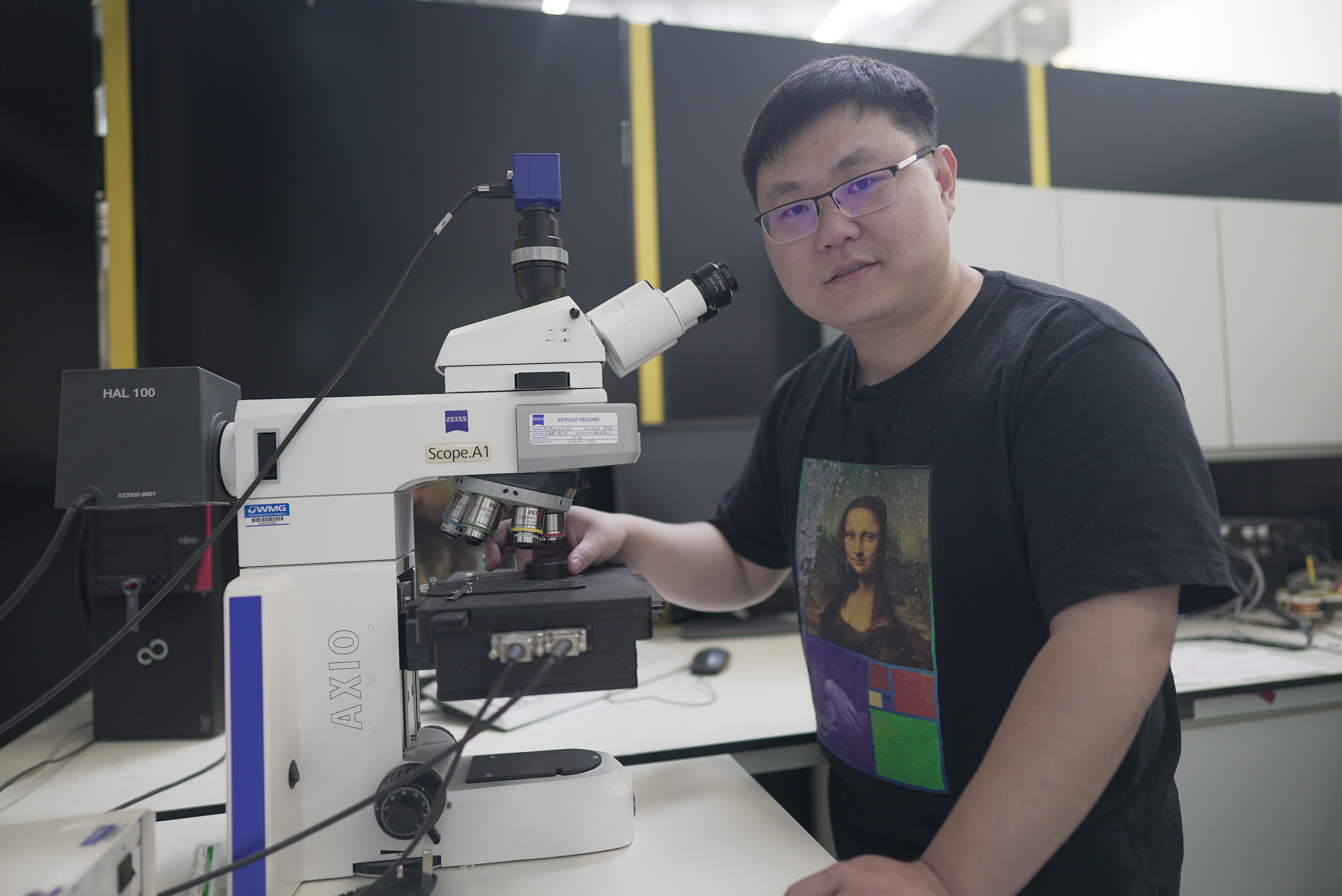 Dr Fanfu Wu standing in a research lab. On the the table next to him is a microscope which is he touching with one hand. 