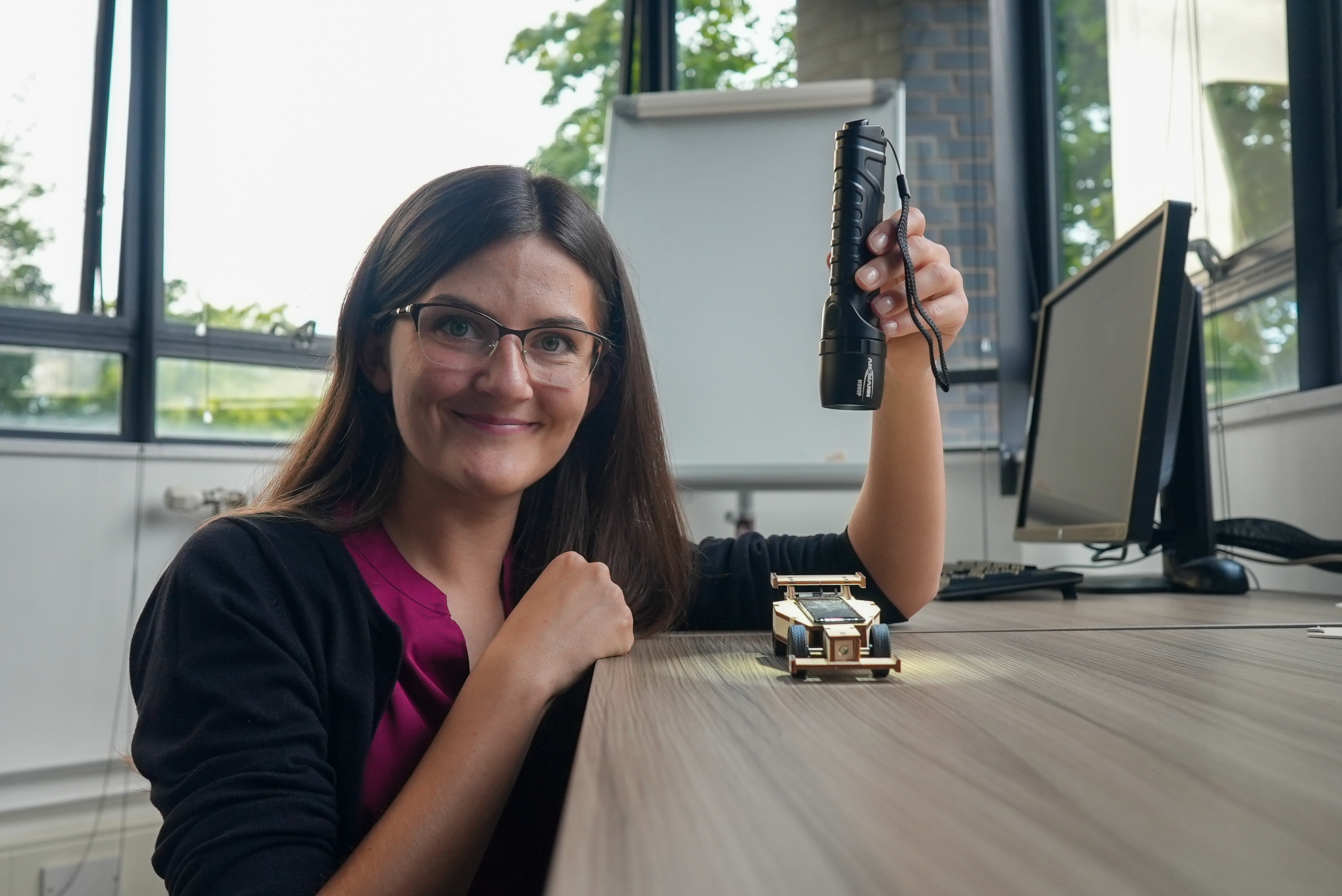 Dr Magdalena Cieslak crouching next to the side of a table holding a lamp above a toy solared power car that is on the table