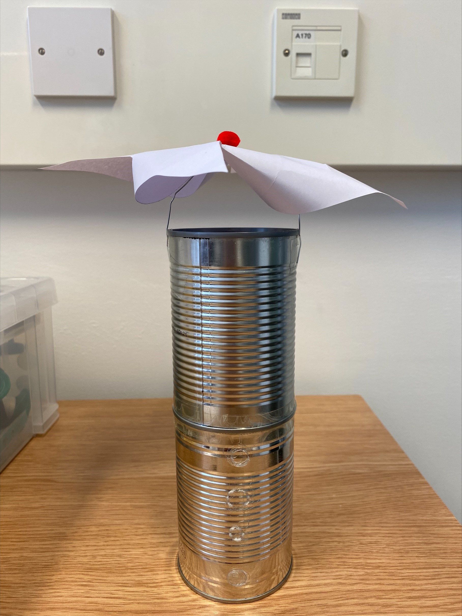 Two tin cans attached one on top of each other. There is a paper clip across the top and a paper windmill on top with a pom pom.