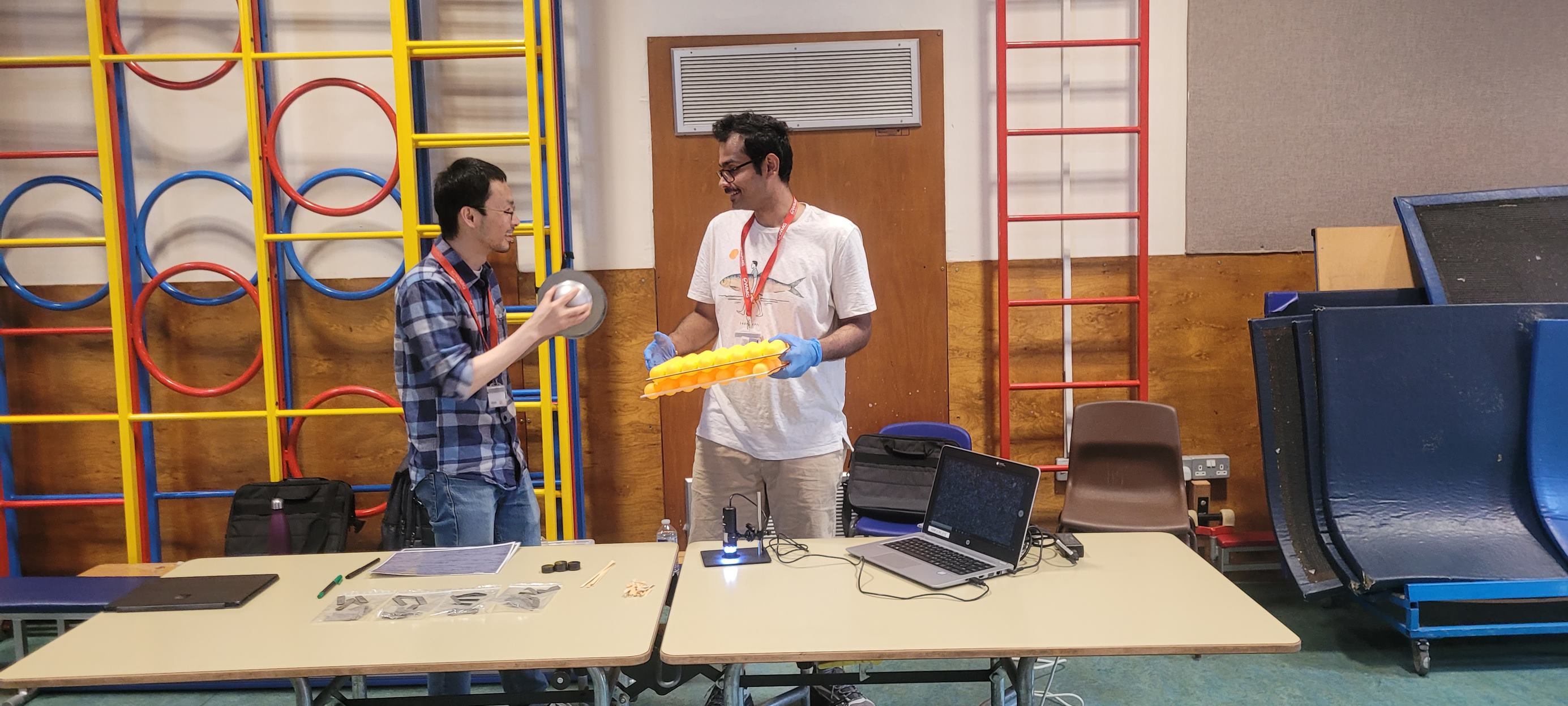 Two people in a school hall.  One person is holding an experiment and showing it to the other person. 