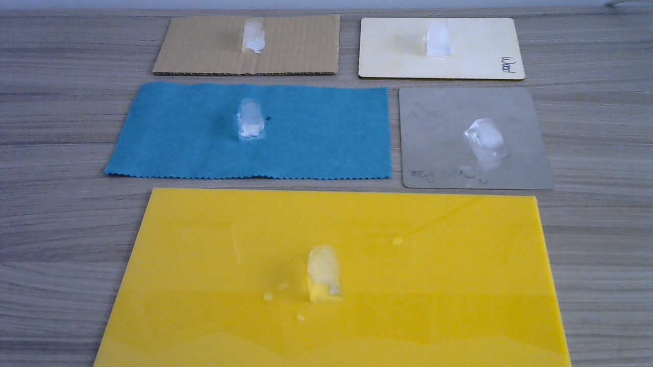 5 rectangular materials placed on a table: felt, cardboard, plastic, metal and  wood. There is an ice cube on each.