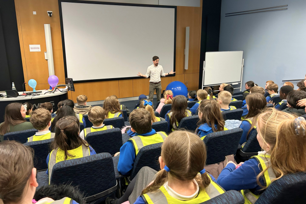 A man presents a science demonstration to an audience of primary school children. 