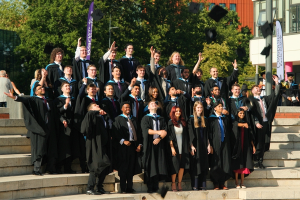 A group of graduates toss their caps in the air while wearing black gowns with sky-blue hoods