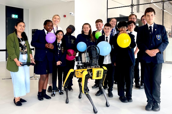 Group of school students holding balloons and smiling with a black and yellow robot dog. 