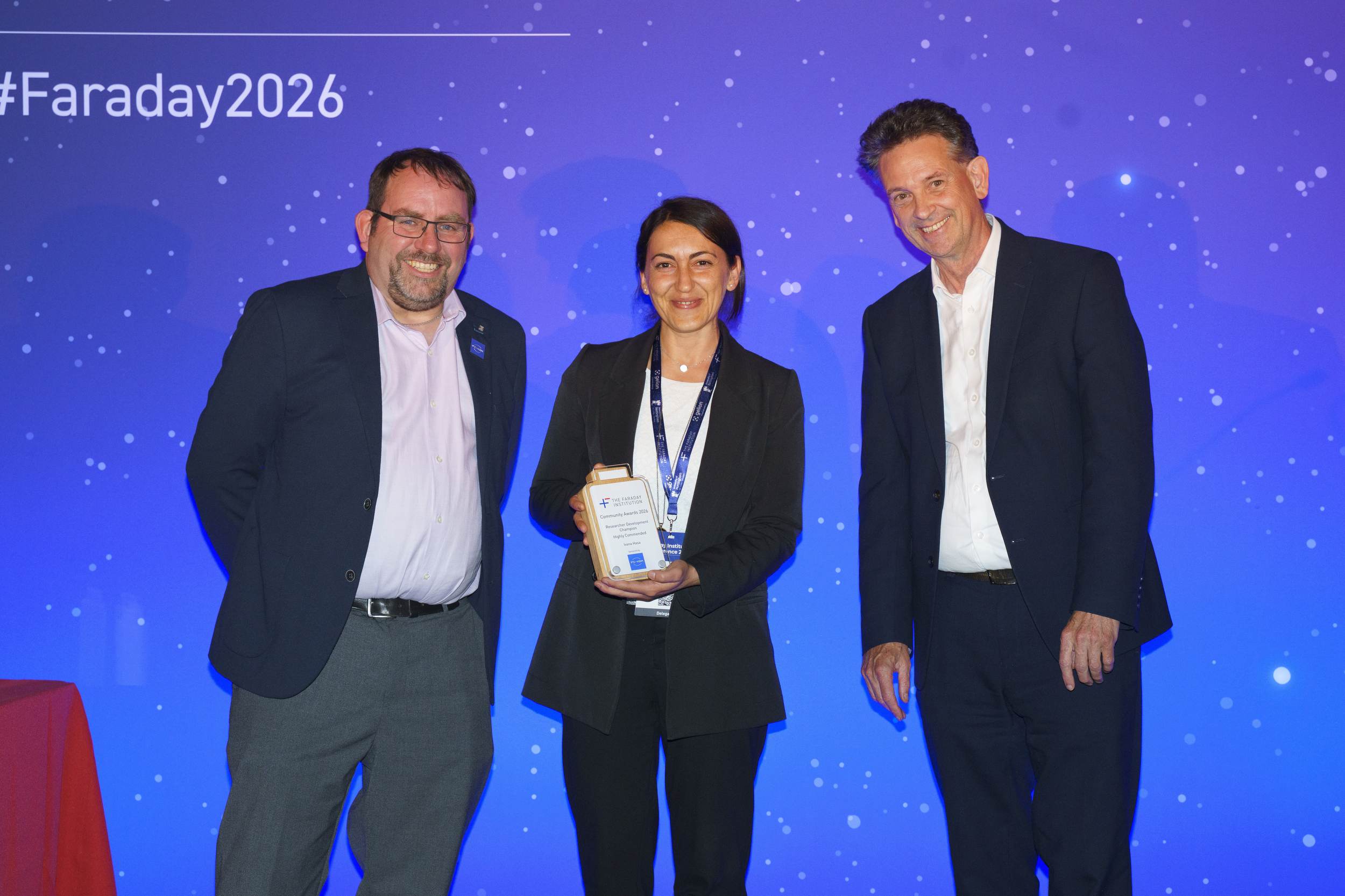 A person stands smiling while holding an award on stage, flanked by two people