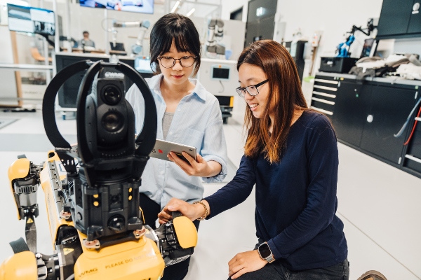 Two female students look at a piece of robotic technology in a lab environment. 