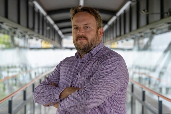 Person standing with arms crossed on a glass-sided bridge, wearing a light purple patterned shirt.