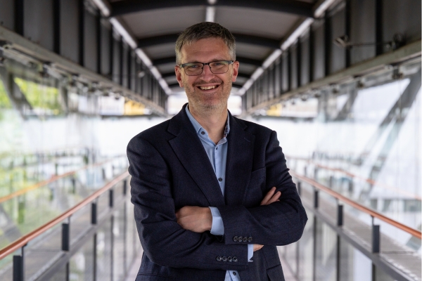 Person wearing glasses, a light blue shirt and a dark blazer, standing with arms crossed on a modern glass-sided bridge.