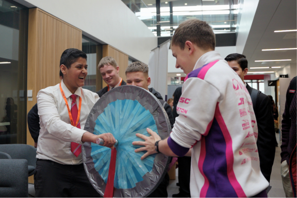 Year 8 and 9 students hold a wheel-like structure and laugh during an engineering demonstration.