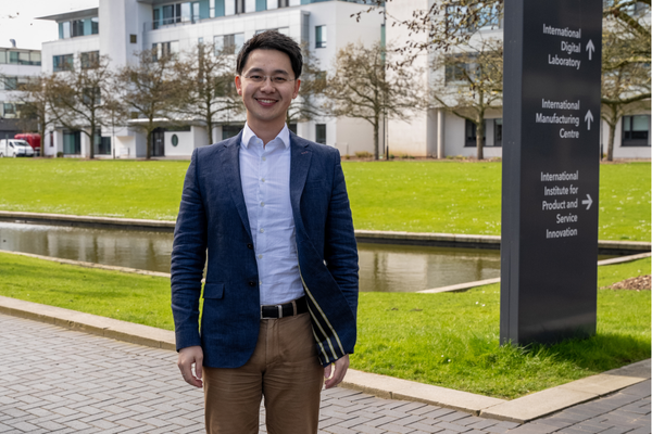 Dr Han Zhang smiles to the camera whilst standing in front of a lake on campus.
