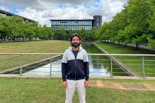 Saif Islam standing outside with greenery and water in background.