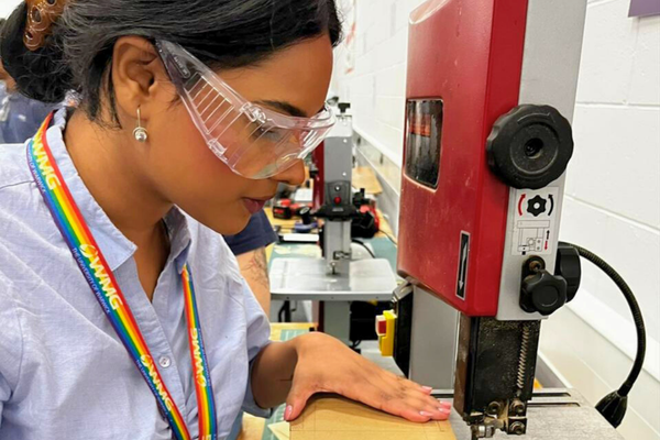 A young woman cuts wood with a machine, whilst wearing safety goggles.