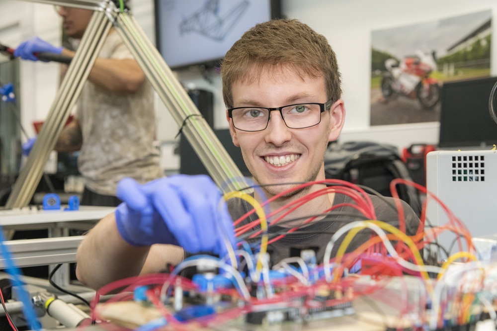 Student working on a robotics project, smiling at the camera while holding wires in place