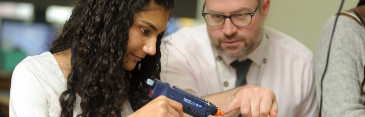 Teacher and student in a design class with a glue gun in the student's hand 