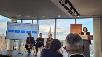 Photograph of the panel at the Modelling an Evolving Economy workshop. Panel consists of 2 white middle men and 2 white middle aged women, sat on a stage beside a lectern and a screen, with a glass window and view of St. Pancras Station behind