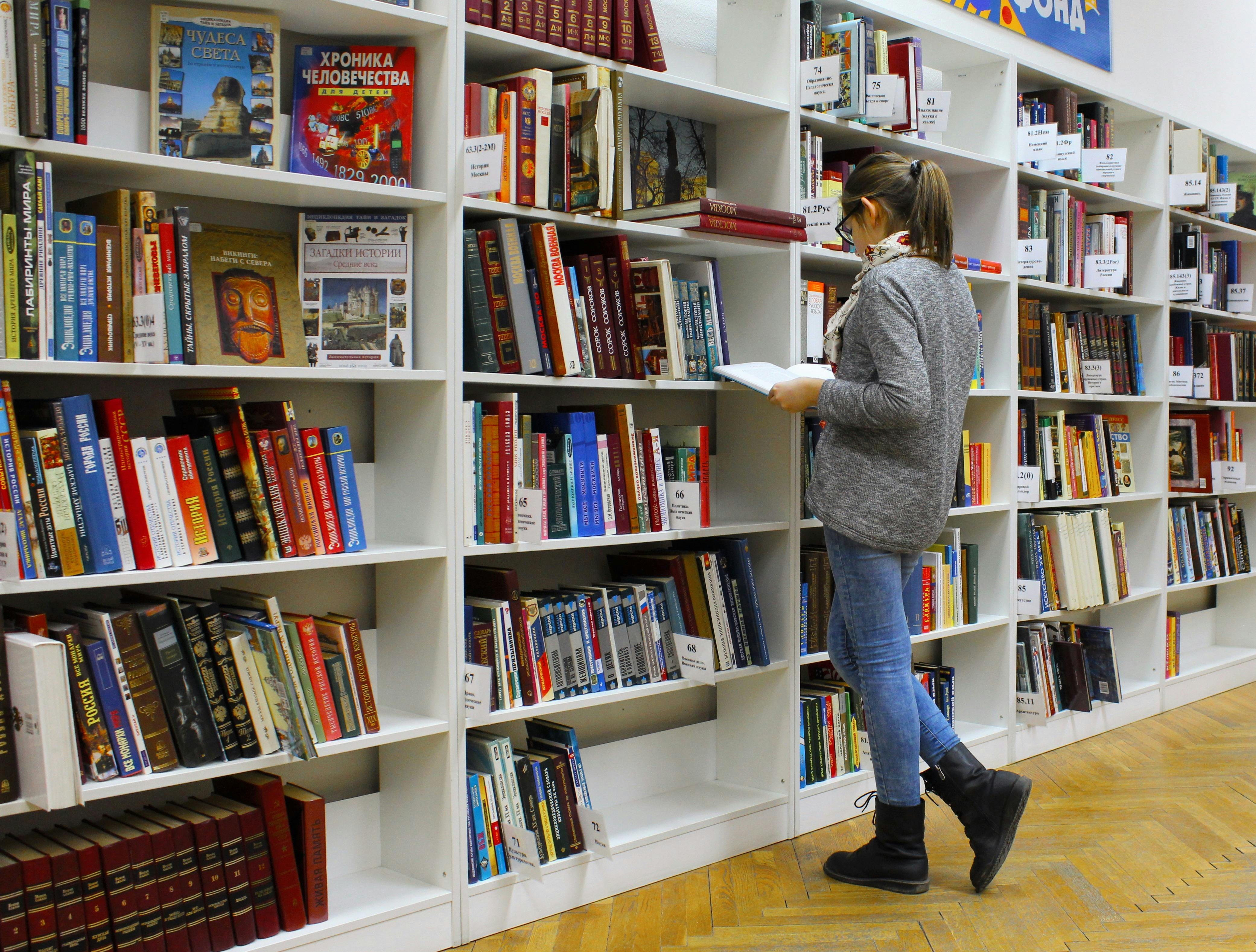 In a library, a person looks at a book in front of a large bookshelf