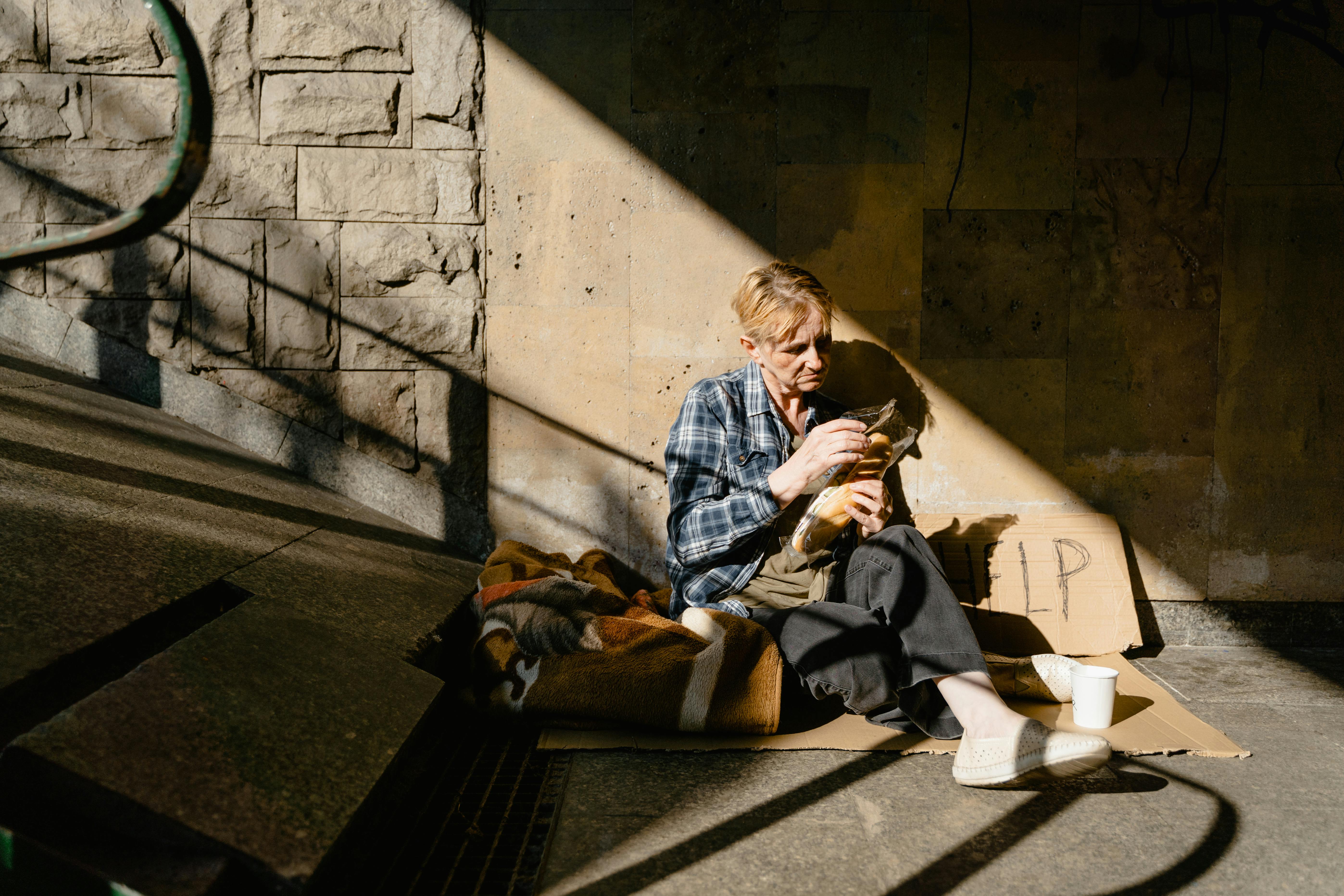 An unhoused person sits at the bottom of some stairs with a 'help' sign and blankets next to her