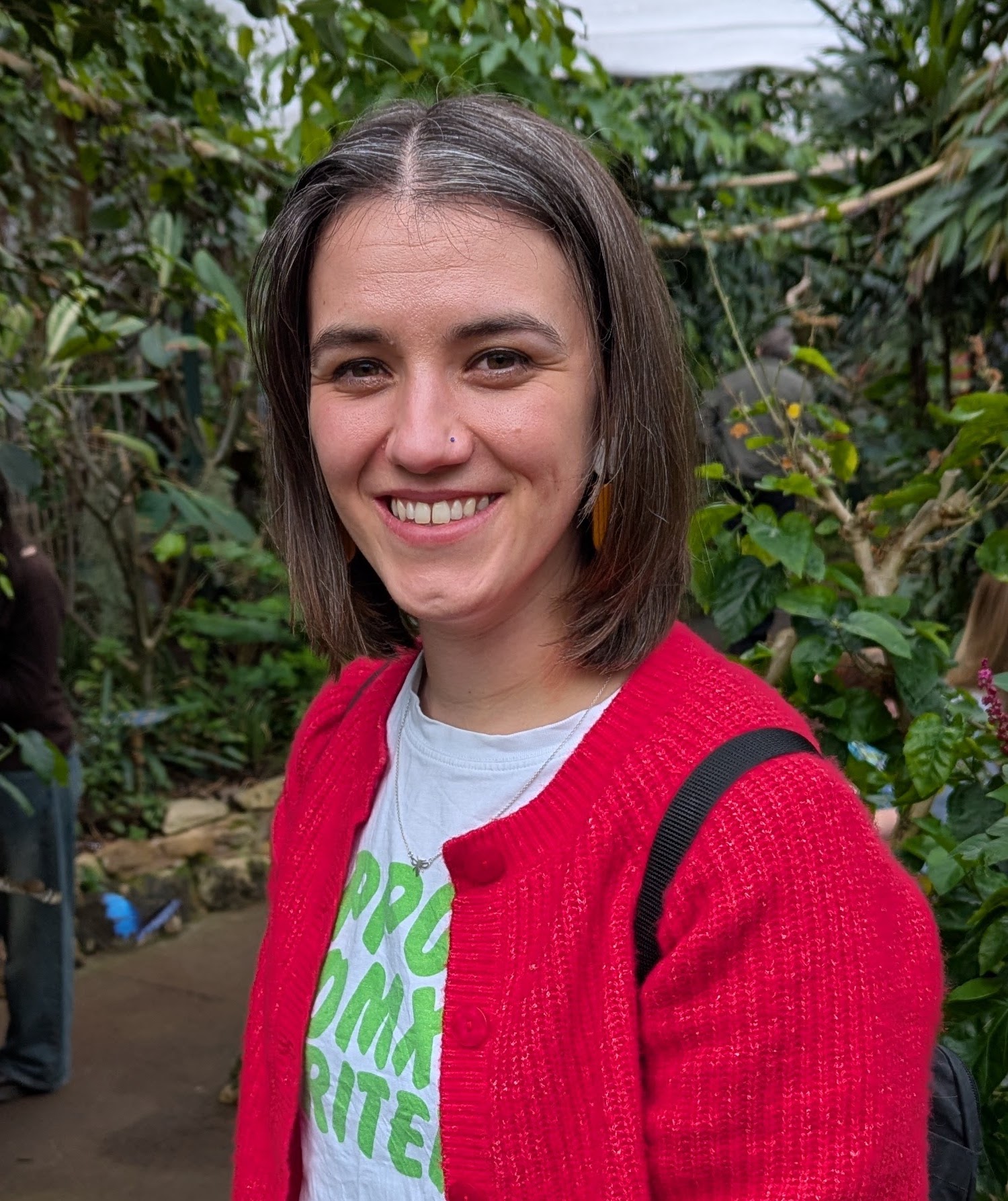 A white woman is standing in front of a background of plants. She is smiling and looking straight at the camera. She has brown hair and is wearing a red cardigan
