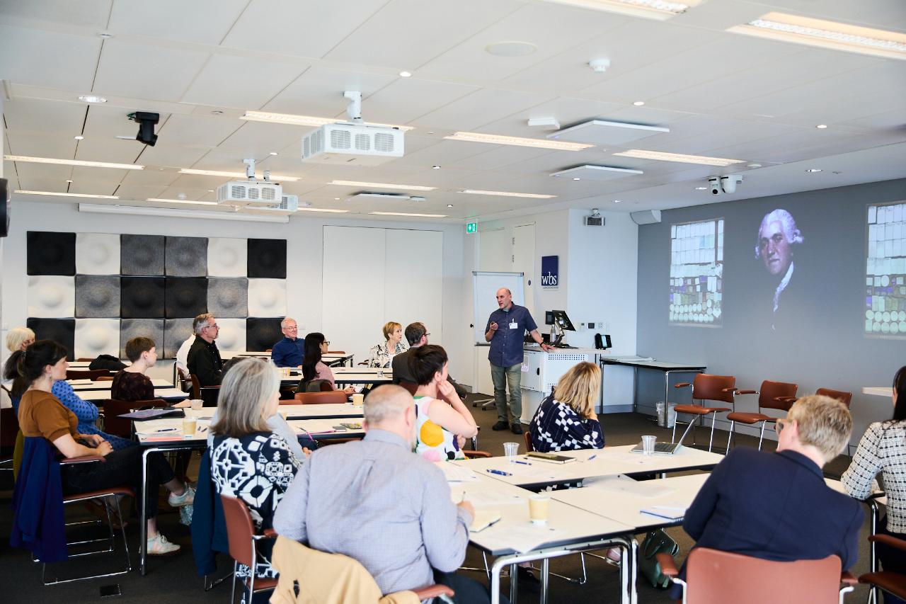A presenting to a room of adults, the slideshow has a portrait of Josiah Wedgewood 