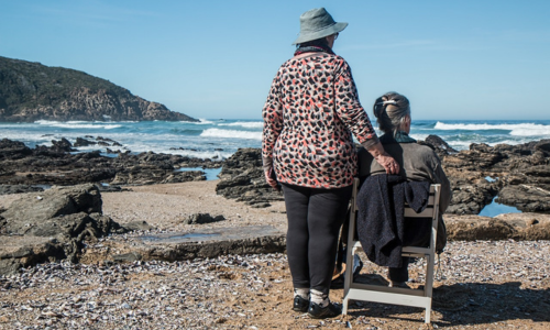 Image of woman seated and woman standing with her arm around the first woman. They are on a beach on a sunny day with a blue sky.