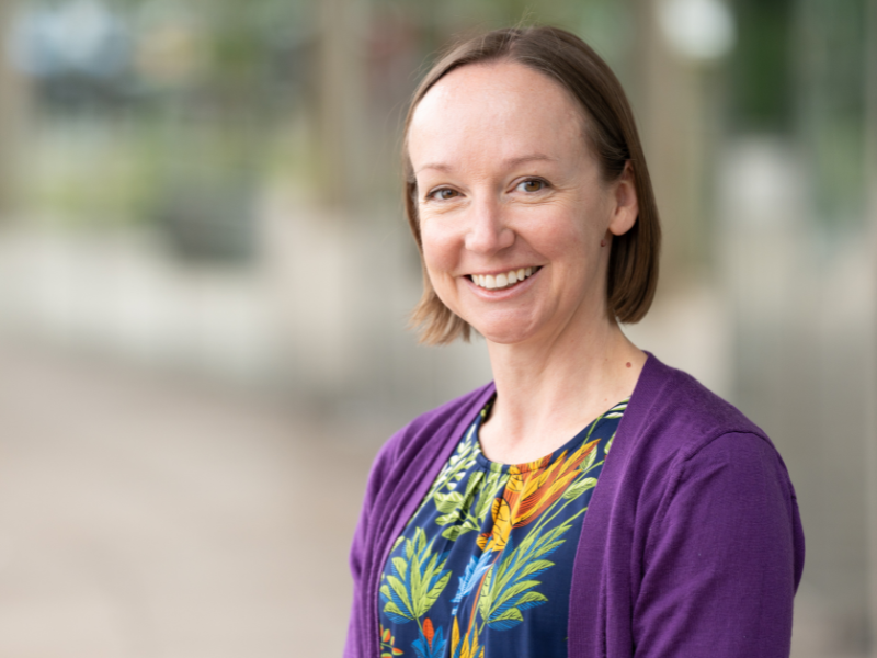 Image of Sarah - a white woman with short brown hair - smiling. She is wearing a bright multicoloured shirt and a purple cardigan. 