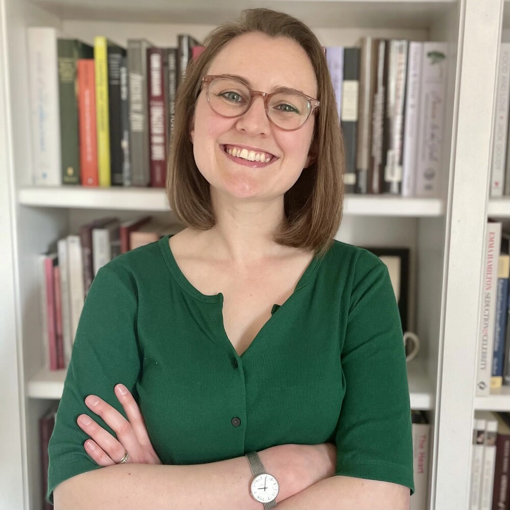 A headshot of Dr Sally Holloway wearing a green top stood in front of shelves of books