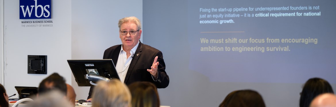 A presenter at the event speaking to the room with a screen in the background