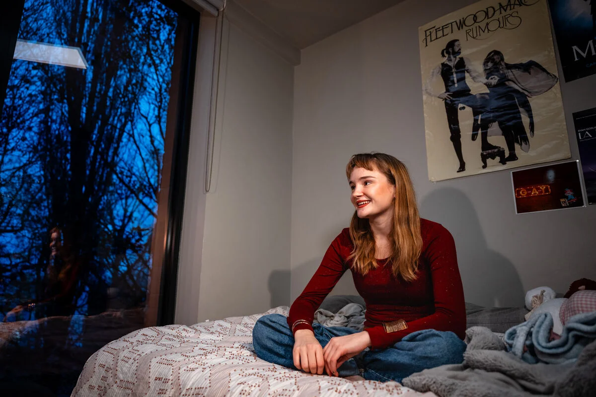 A student in a red top is sitting on a bed in a campus room decorated with posters on the wall 