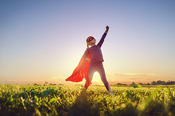 A child strikes a superhero pose, dressed in full superhero costume, in a field, as the sun sets behind them