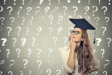 A young woman wearing a graduation hat, pondering with her finger to her lips, sitting in front of a wall of question marks