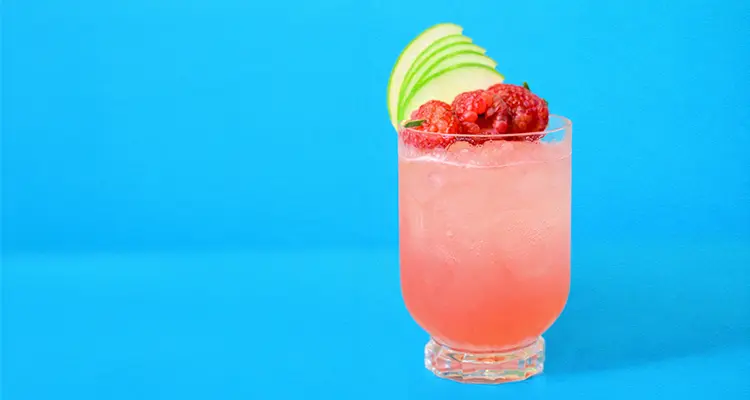 Pink cocktail in a glass with raspberries and apple garnish at the top of the glass