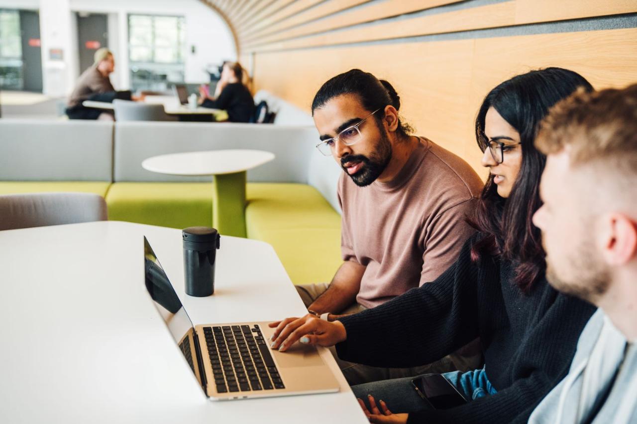 A group of people sit together at a table in a modern study area, looking at a laptop on the table.
