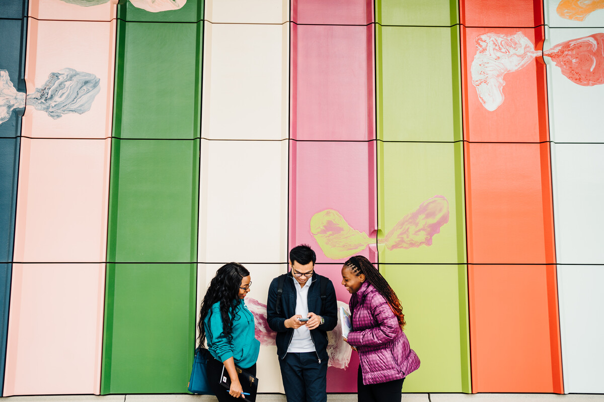 Three students standing in front of a colourful background, the middle person is holding a mobile telephone and they are all looking down at the screen