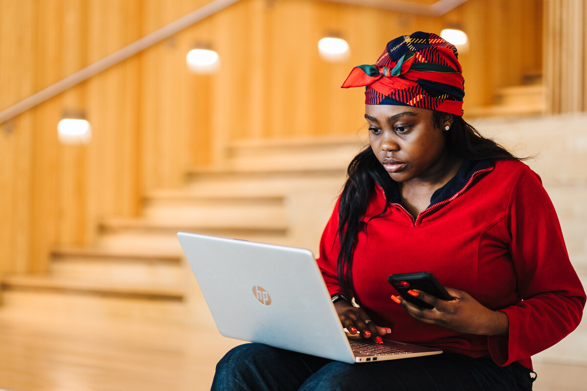 A student sitting on the steps in the Faculty of Art Building, looking at their laptop and holding a mobile telephone