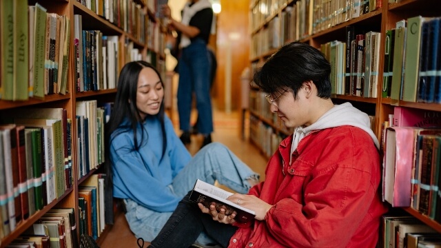 Male student in red jacket sitting on the floor beside female student in blue long sleeve shirt