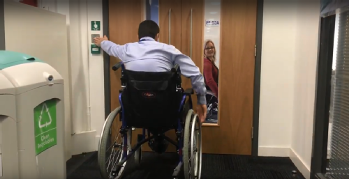 A participant of the wheelchair challenge presses the "open door" button before trying to open the double pull doors. Another participant watches through the glass panes in the doors.