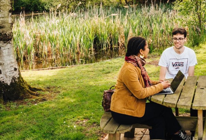 Students on a bench with laptops in green surroundings next to a lake