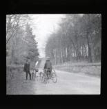 Men on tricycles on country road, bridge and trees in background