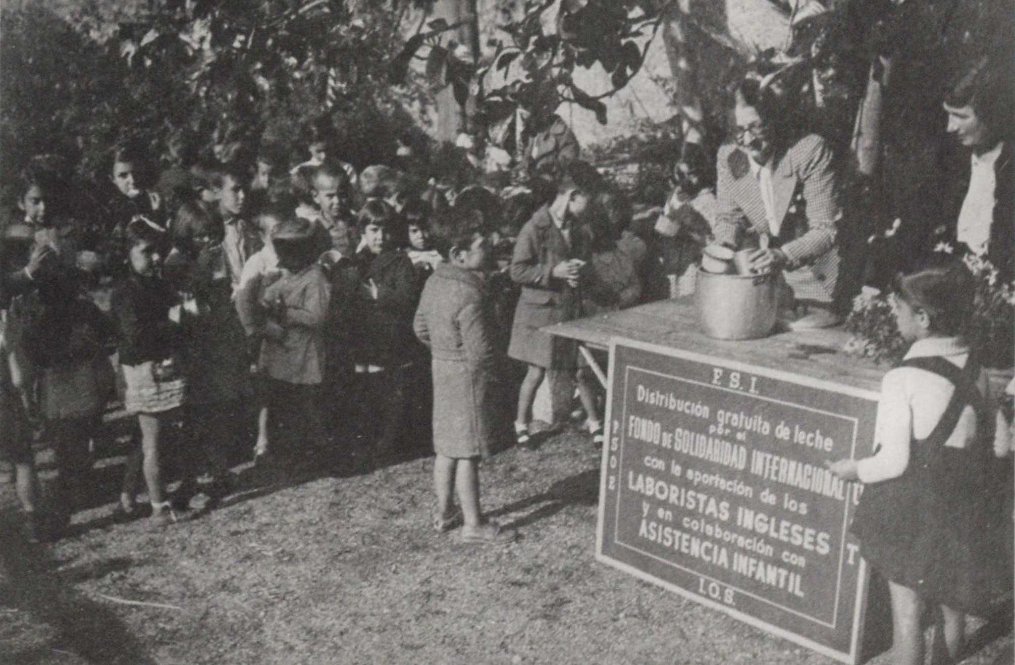 Woman standing behind a table and pouring milk into mugs for a group of children gathered around. 