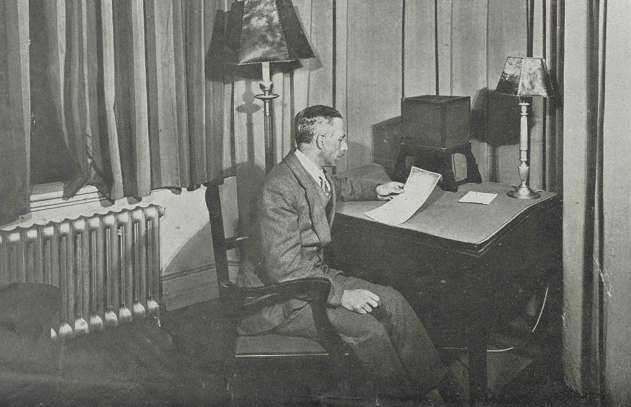 BBC announcer at a desk. He is holding a piece of paper.
