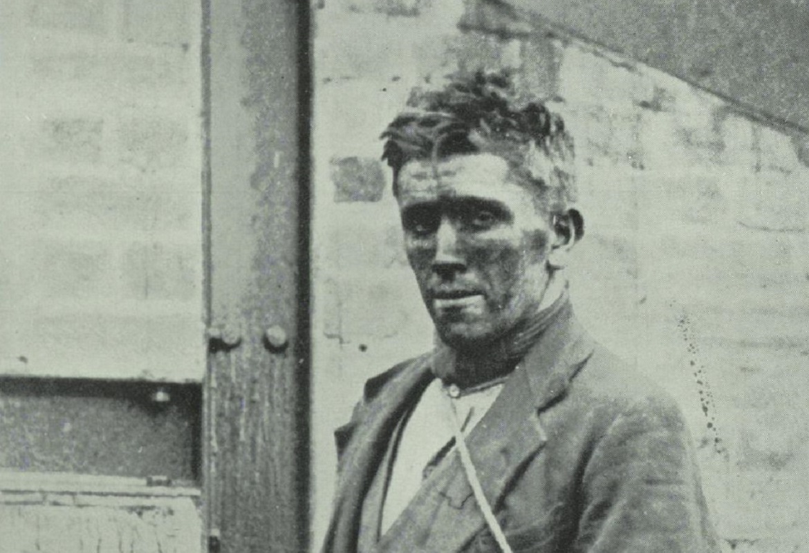 Black and white photograph of a miner. He is looking towards the camera and has a face covered with coal dust after coming up from the pit.