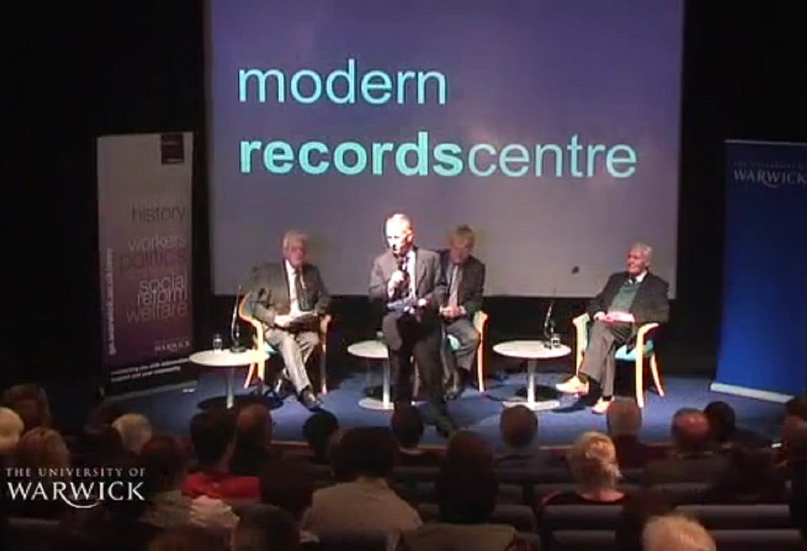Three men seated on a stage, with a fourth man standing in front of them. The name of the Modern Records Centre is on a screen behind them.