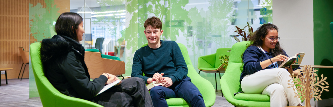 Three people sat on green chairs. Two are facing each other and smiling, a third is facing a different way, reading a book and smiling at it.