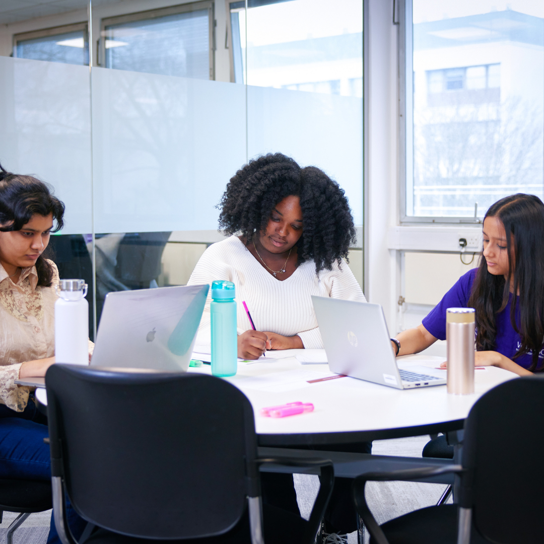 Two students working, one on a laptop, one writing, next to a Library Student Partner who is also working beside them. A depiction of our Write Here Write Now event.