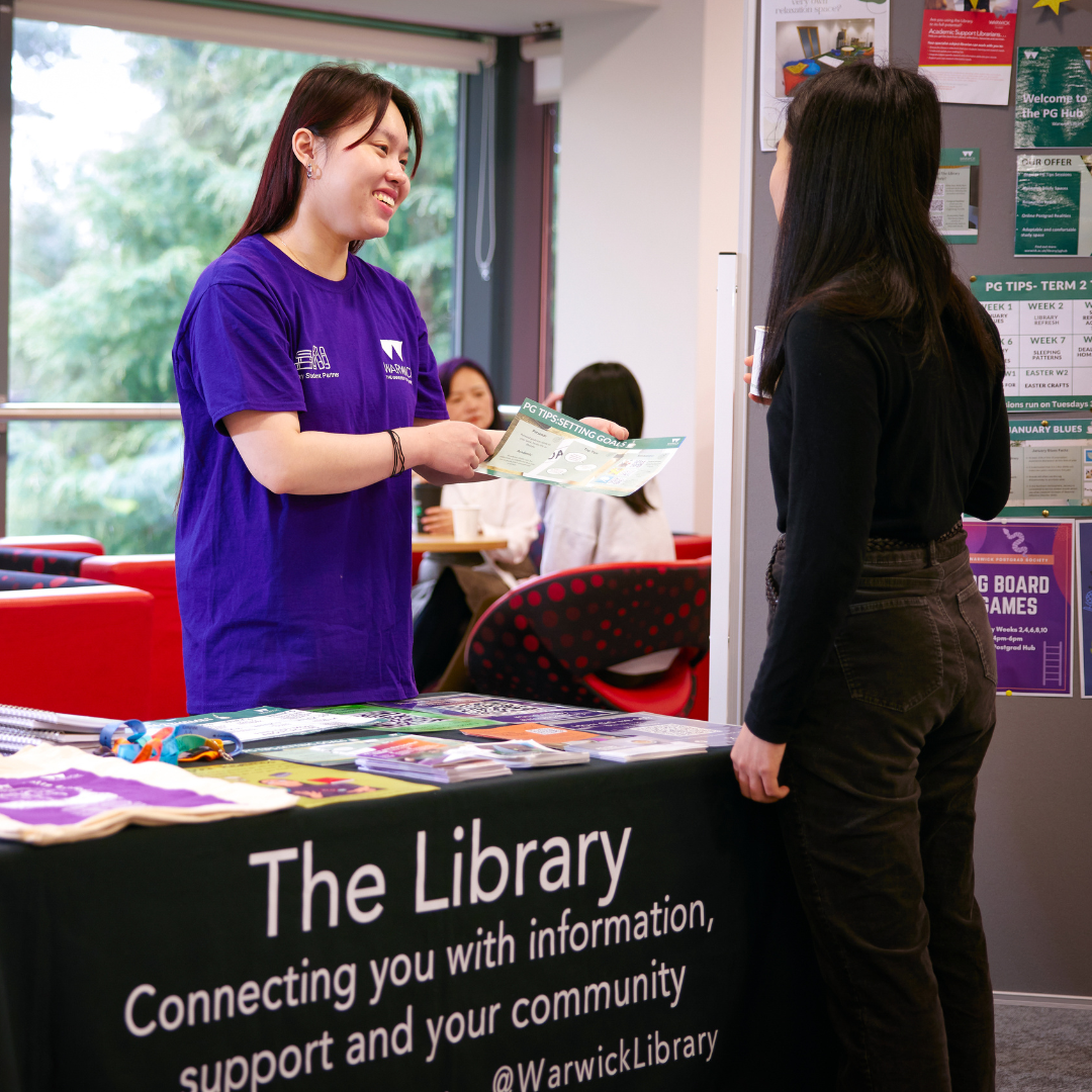 A Library Student Partner holding out an information page and helping a student at a stand. The Library is prominently displayed on the table.