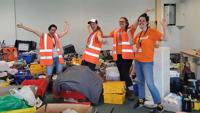 Four people wearing orange hi vis jackets are standing in a room full of stuff.