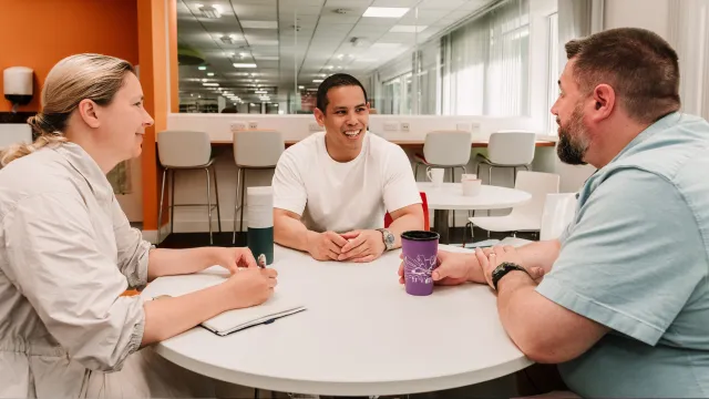 Three people are sitting around a table in a office staff kitchen.