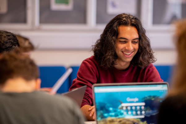 A group of students are sait working at their laptops, one is facing the camera and smiling.