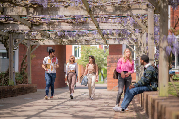 A group of students walk and talk under a wisteria covered pergola at the University of Warwick