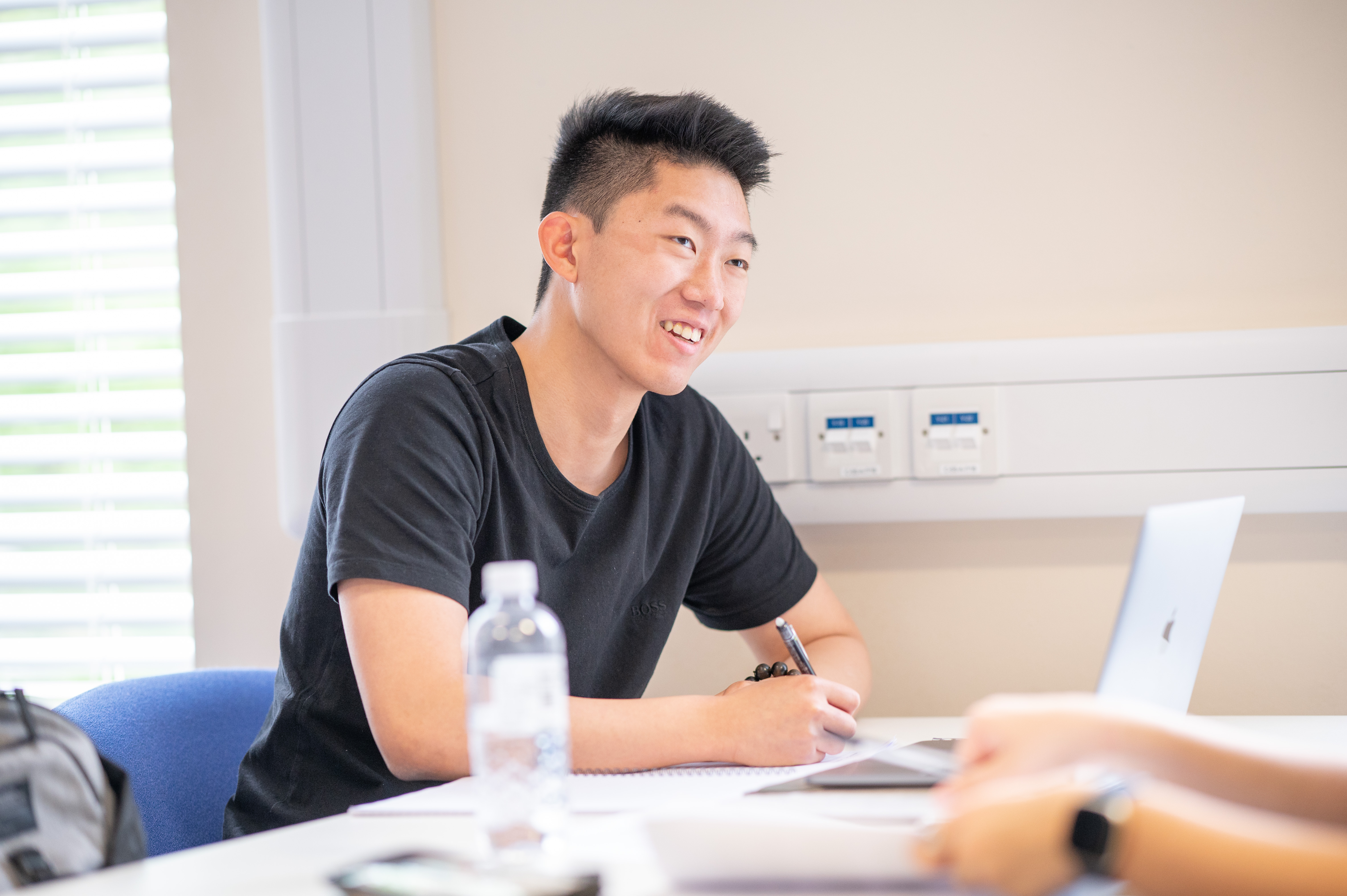 A student smiles as they explain their research to another person who is sitting opposite them at a table